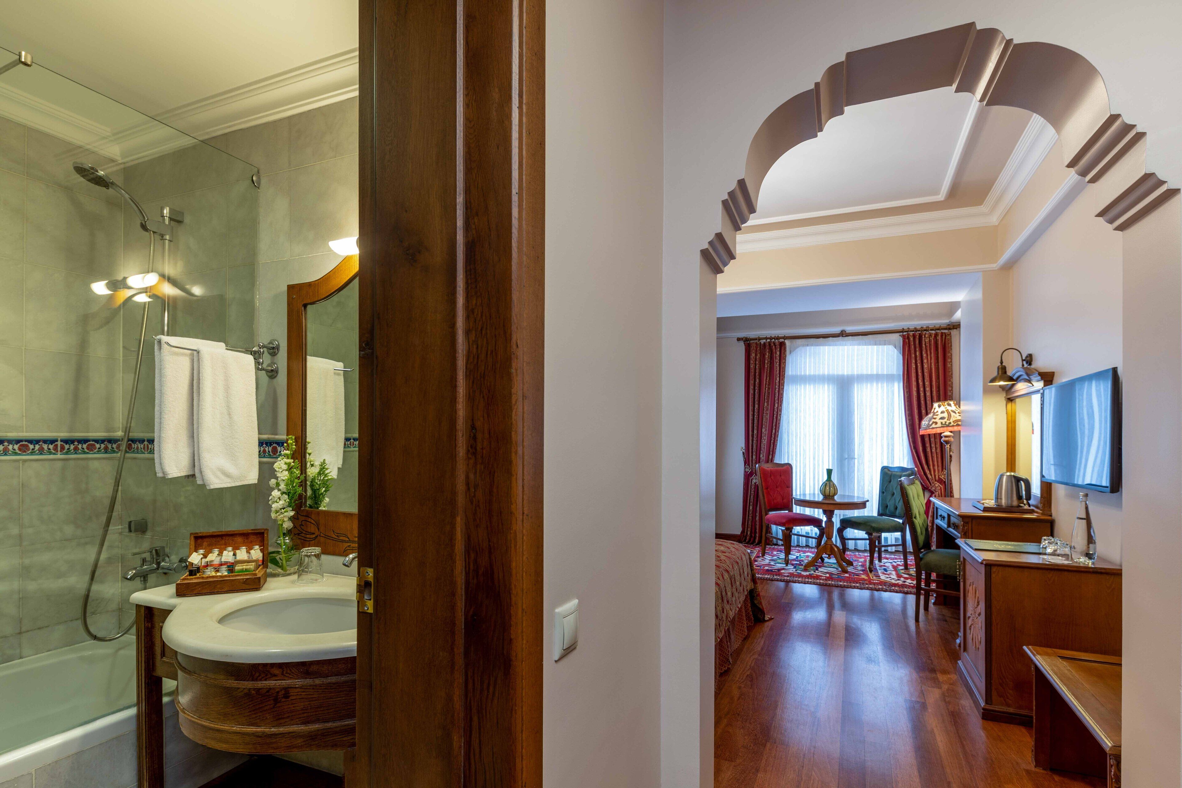 A hotel bathroom with a bathtub, sink, and mirror. There is a wooden vanity and a shower area with a glass door. The bathroom has a tiled floor and a wooden door.