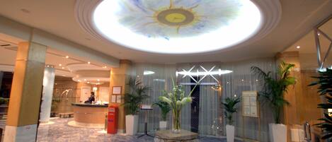 Un vestíbulo de hotel con una fuente decorativa en el centro, un mostrador de recepción y un amplio tragaluz.
