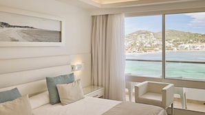 A hotel room with a bed, a desk, and a chair. There is a large window with a view of a beach and buildings. - Argos Hotel (Ibiza Town)