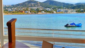 A wooden table with chairs in a room with a large window overlooking a body of water, a boat, and a distant hillside. - Argos Hotel (Ibiza Town)
