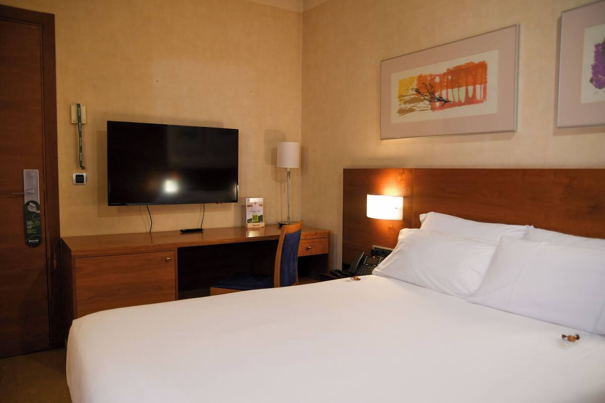 A hotel room with a large bed, a desk with a flat-screen TV, a telephone, and a wall-mounted lamp.