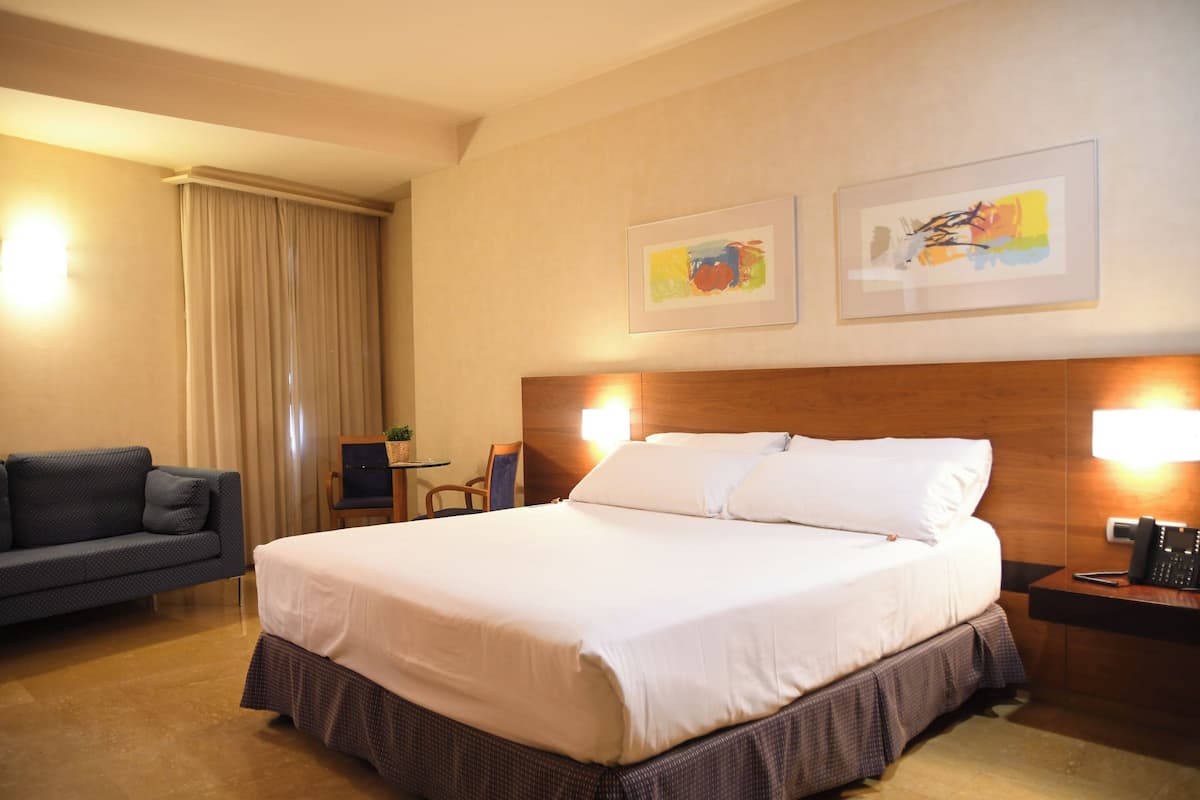 A hotel room with a large bed, a sofa, two framed artworks on the wall, and a desk with a lamp.