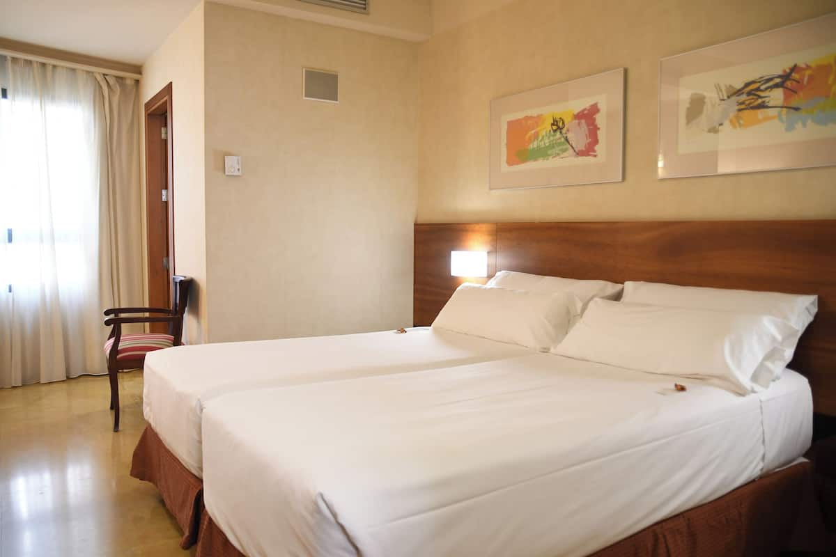 A hotel room with two beds, a wooden headboard, a chair, and two framed artworks on the wall.