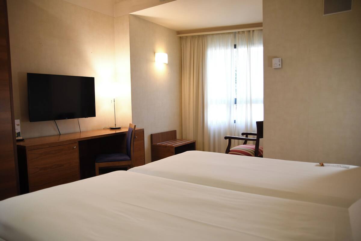 A hotel room with two beds, a desk, a chair, a TV, and a window with curtains.