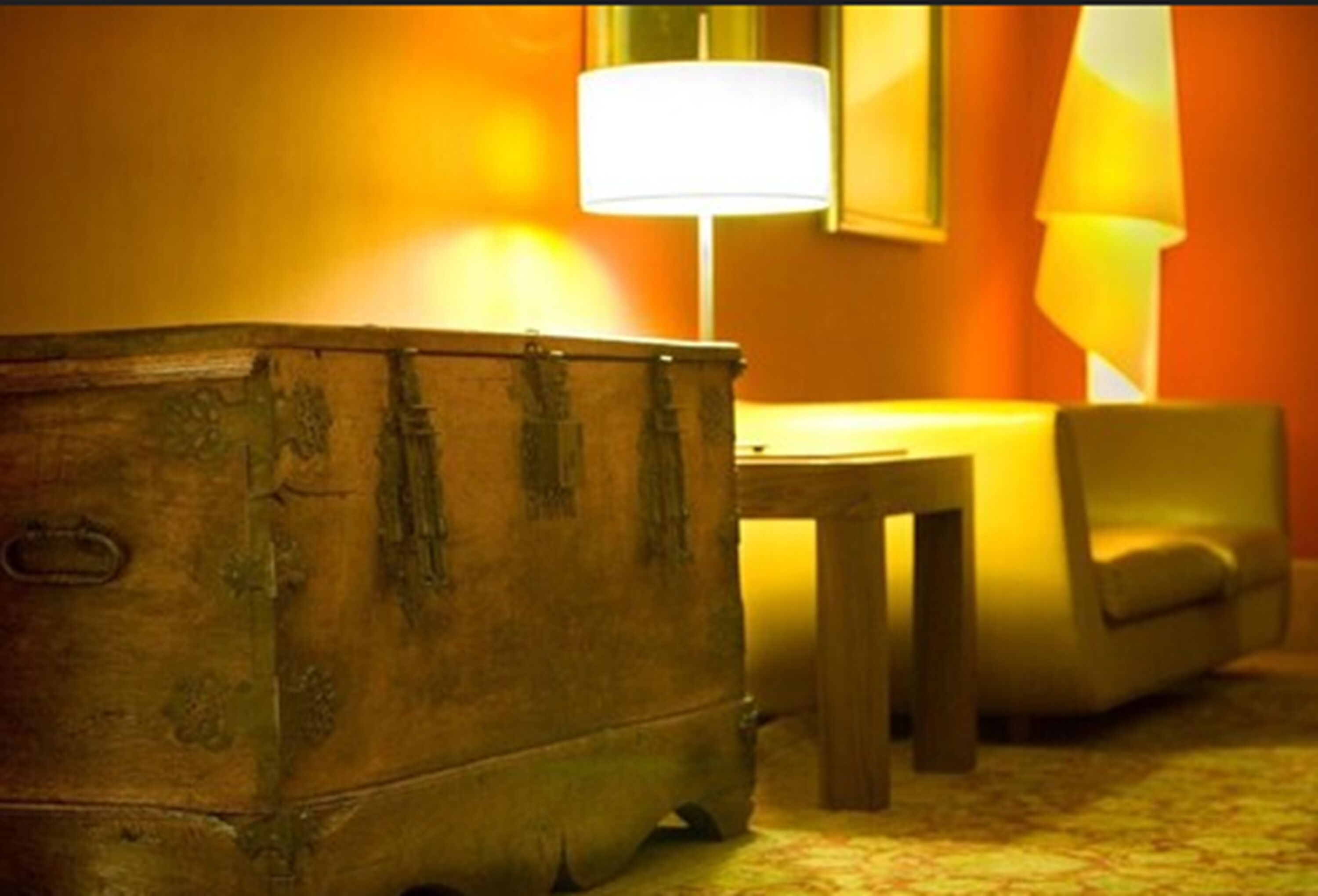 Un cofre de madera en una habitación cálidamente iluminada, con una lámpara y una silla.