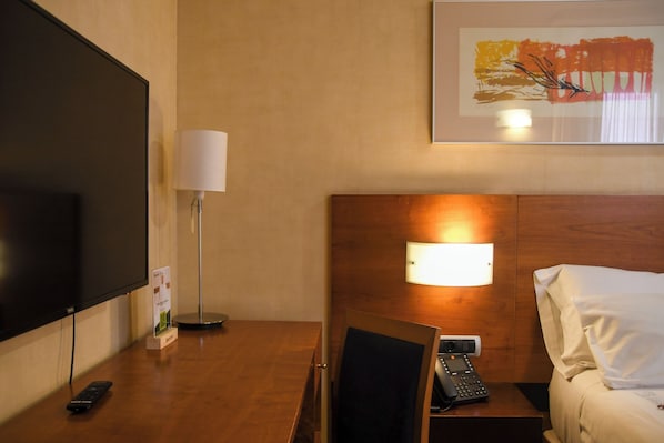 A hotel room with a bed, a bedside table with a lamp, a phone, a TV, and a framed picture on the wall.