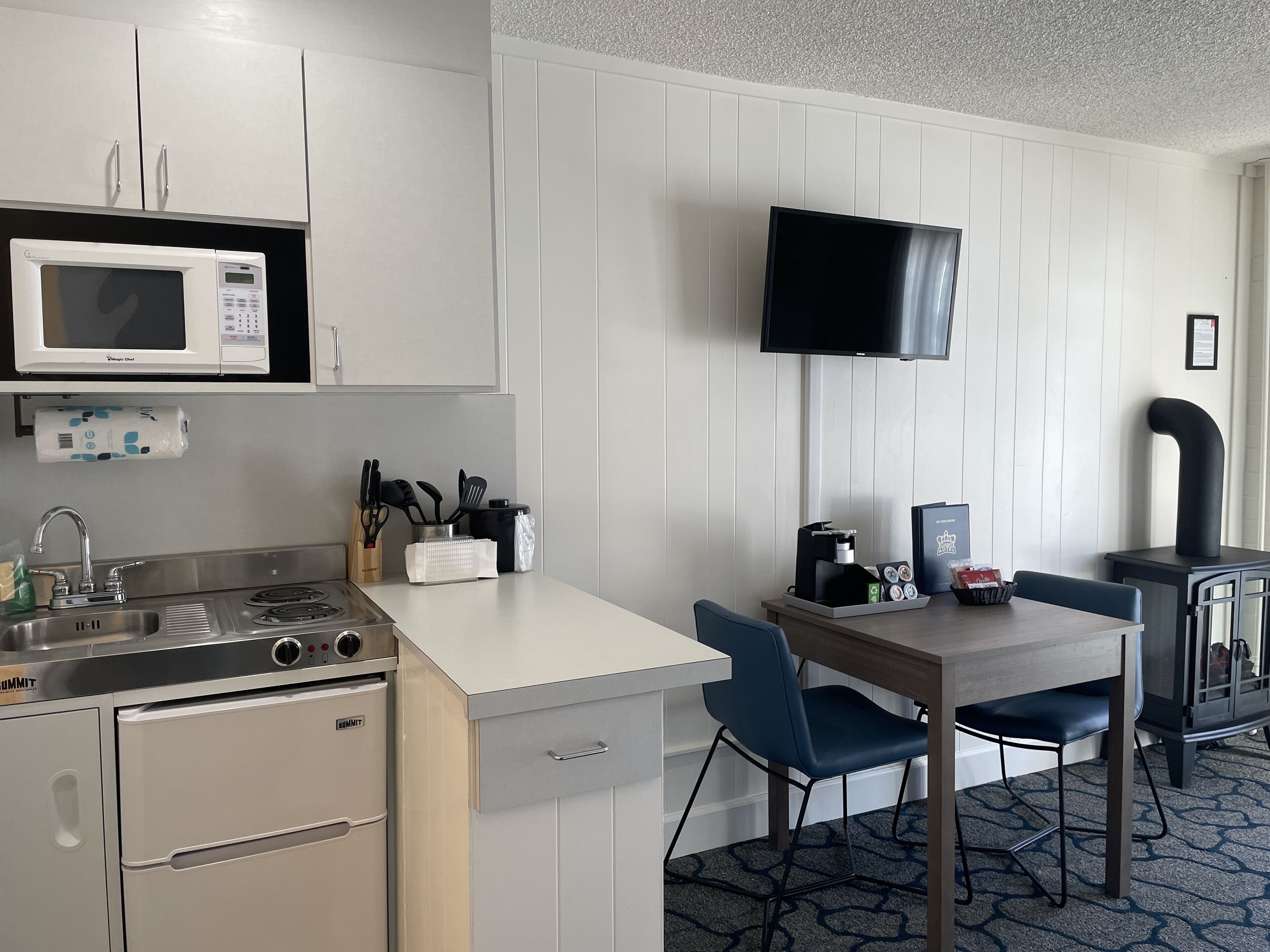 A compact kitchen with a microwave, stove, sink, and a flat-screen TV mounted on the wall.