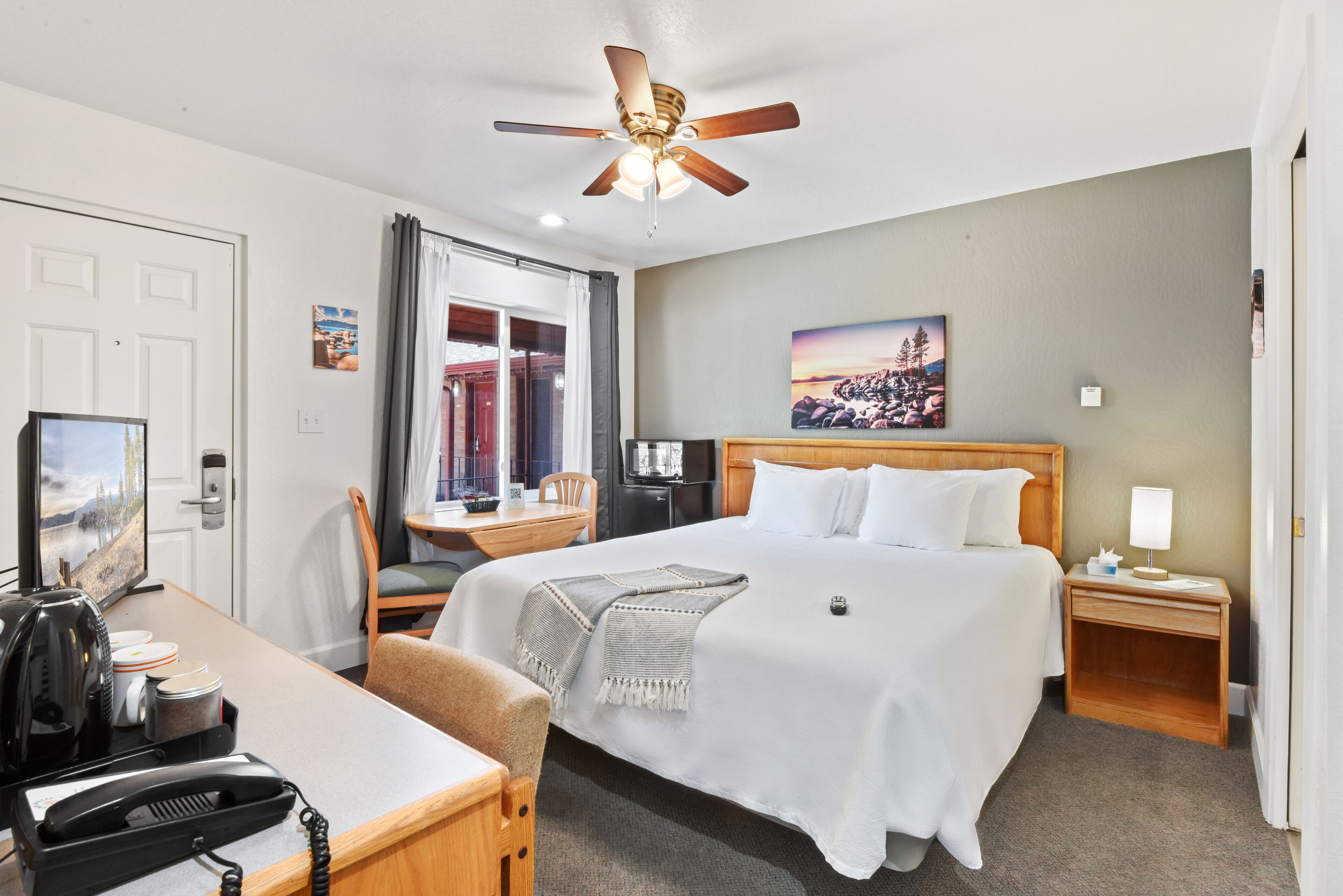 A hotel room with a bed, desk, chair, ceiling fan, and a painting on the wall.