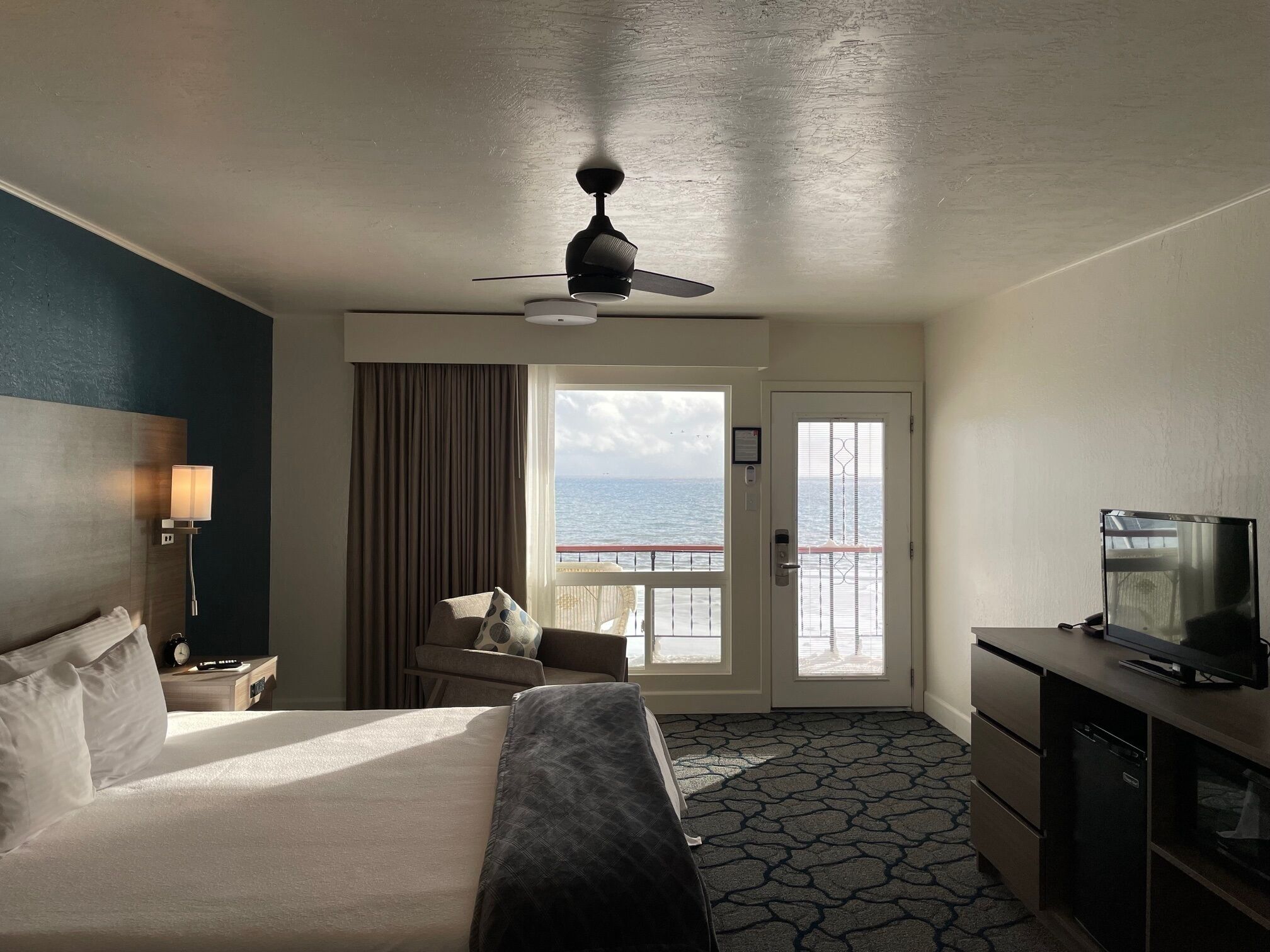 A hotel room with a bed, a desk, a television, a ceiling fan, and a view of the sea through a door.
