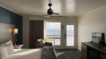 A hotel room with a bed, a desk, a television, a ceiling fan, and a view of the sea through a door.