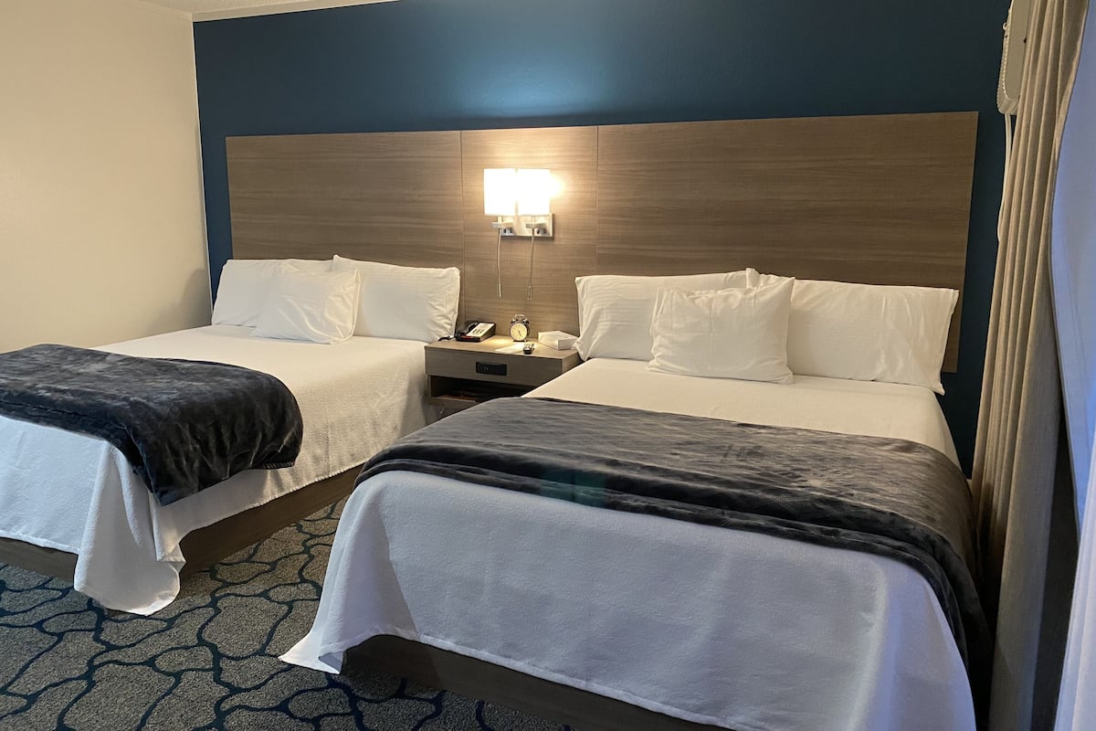 A hotel room with two beds, a nightstand with a lamp, and a wall-mounted light.