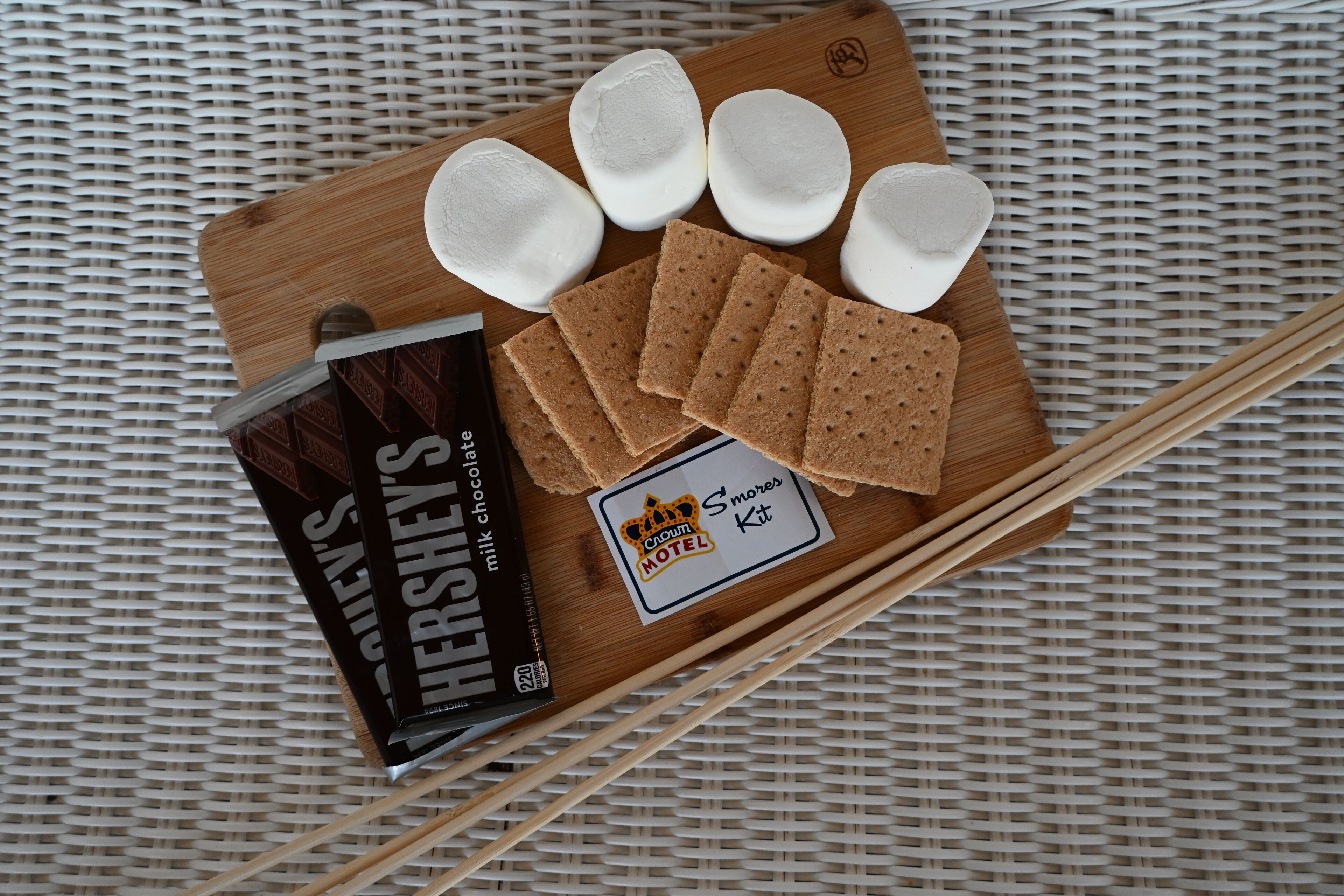 A wooden board with marshmallows, chocolate bars, and a s'mores kit on a woven surface.