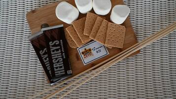 A wooden board with marshmallows, chocolate bars, and a s'mores kit on a woven surface.
