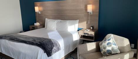 A hotel room with a large bed, two bedside lamps, a nightstand, a chair with a patterned cushion, and a patterned carpet.