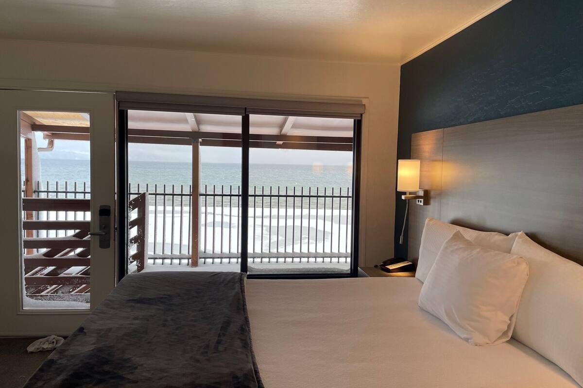 A hotel room with a bed, a nightstand, a lamp, and a view of the ocean through a sliding door.