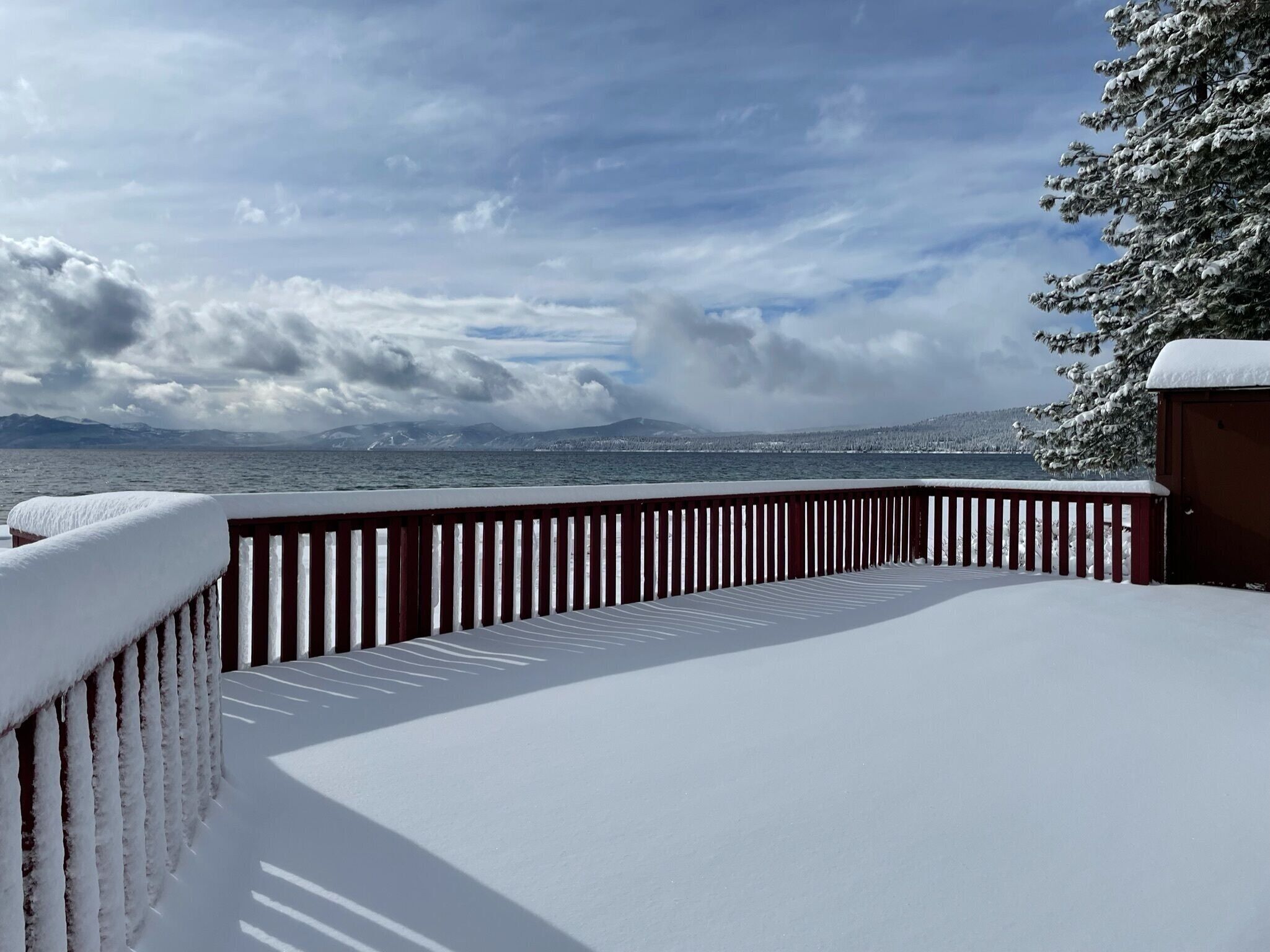 A snow-covered deck with a wooden railing, overlooking a body of water and distant mountains.
