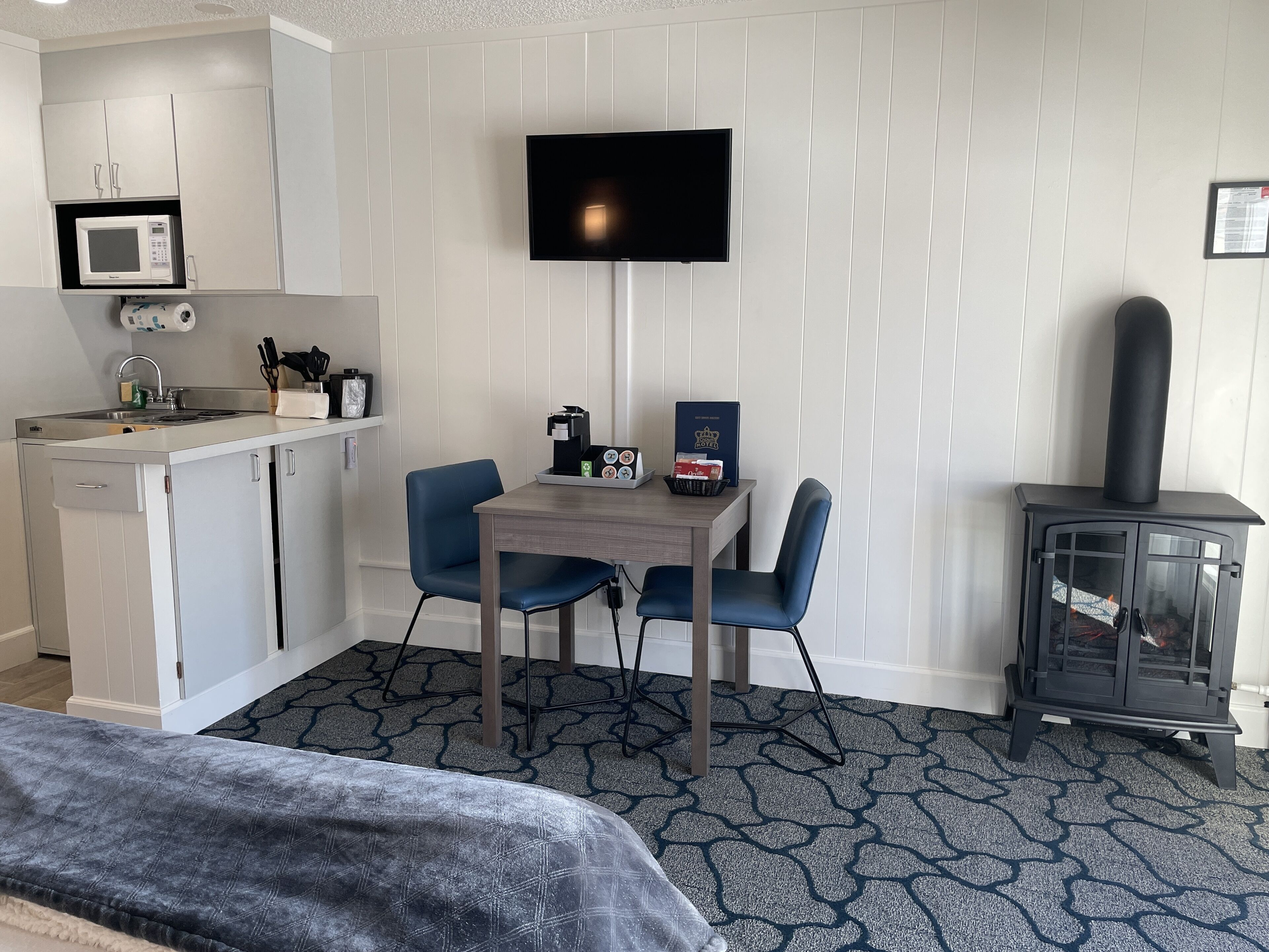 Une chambre d’hôtel compacte comprenant un coin cuisine, un téléviseur à écran plat fixé au mur, une petite table à manger avec des chaises, un lit recouvert d’une housse à motifs gris et un poêle à bois.