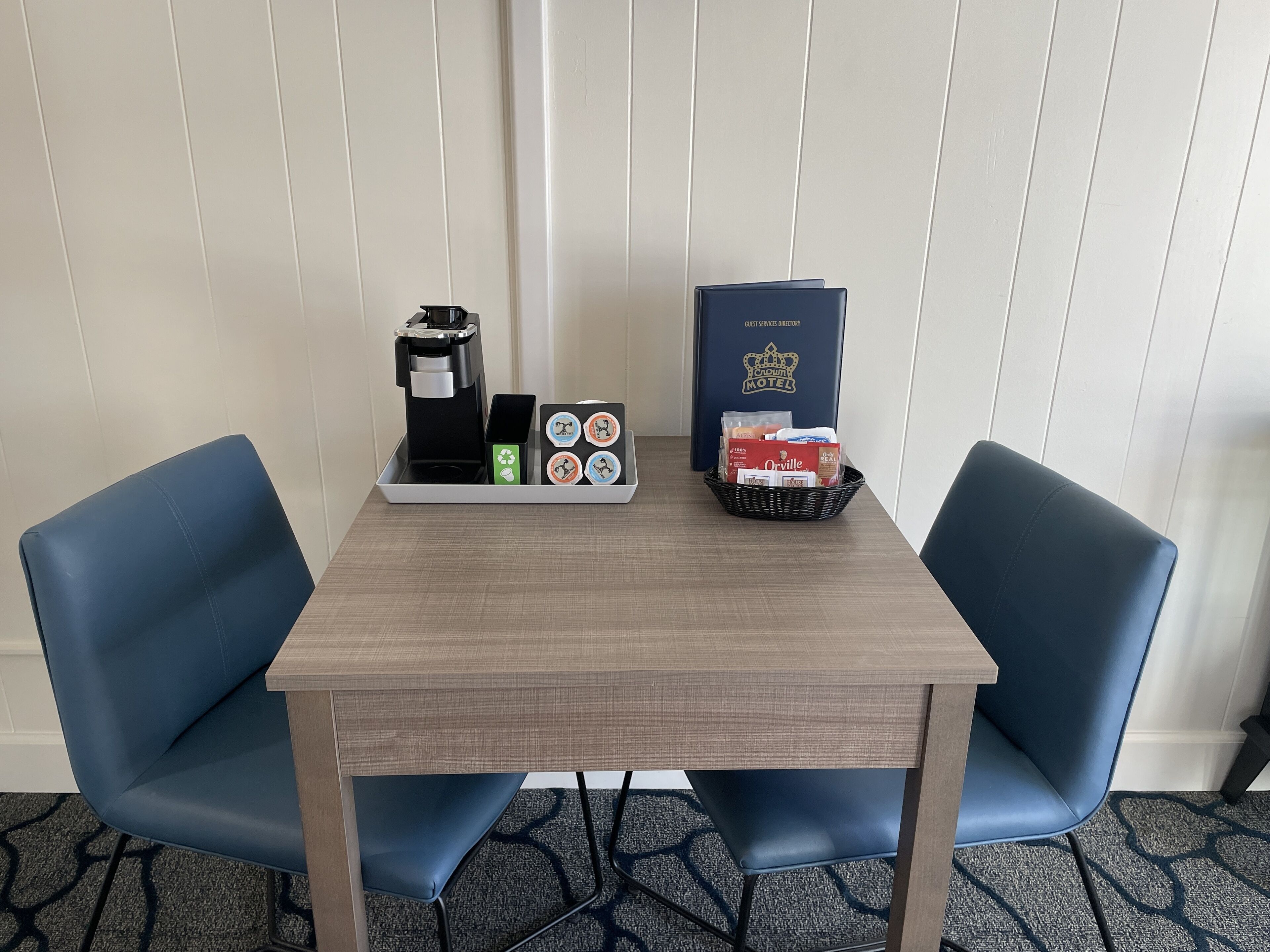 Hotelkamer met een houten tafel, twee blauwe stoelen, een koffiezetapparaat, een gastenmap en een mand met lekkernijen.