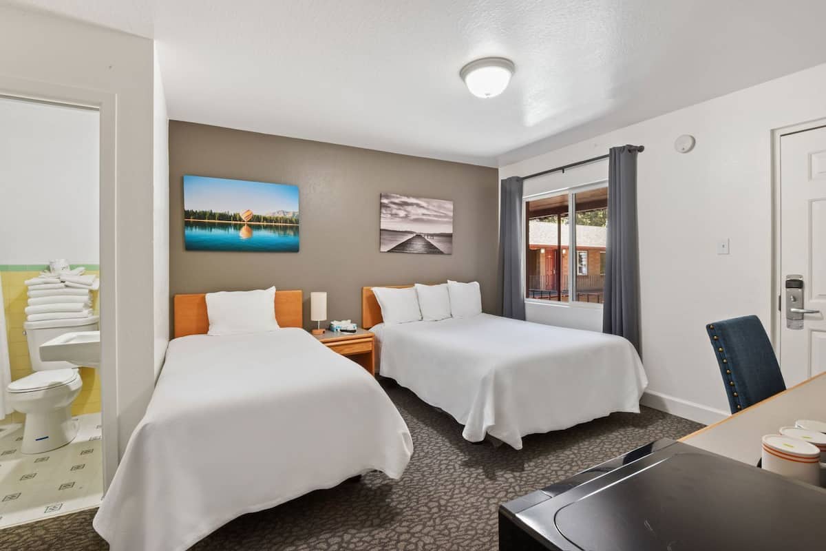 A hotel room with two beds, a desk, a chair, a bathroom with a toilet and sink, and a window with curtains.