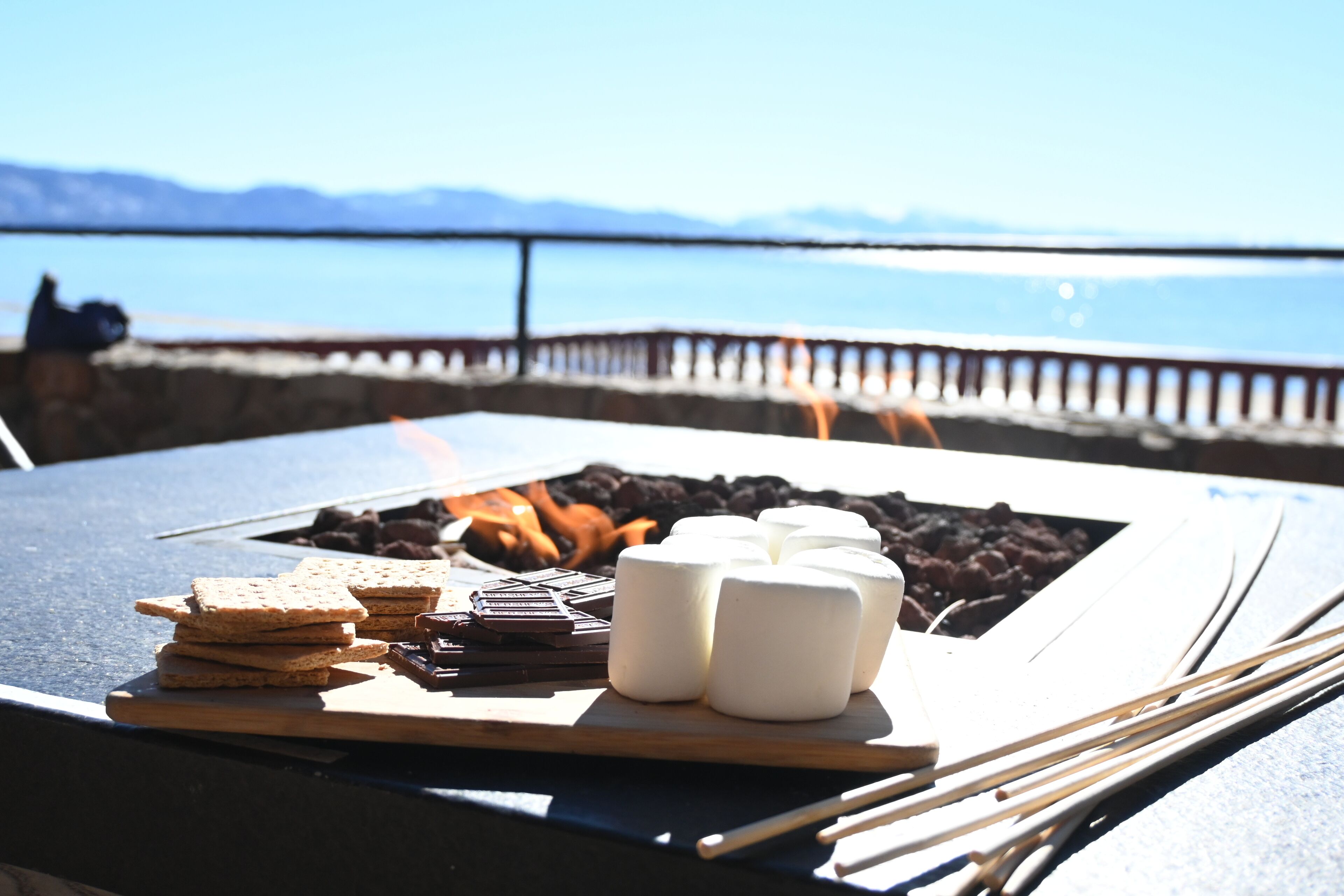 A table with marshmallows, chocolate, and crackers, with a fire pit and a view of the sea.