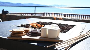 A table with marshmallows, chocolate, and crackers, with a fire pit and a view of the sea.