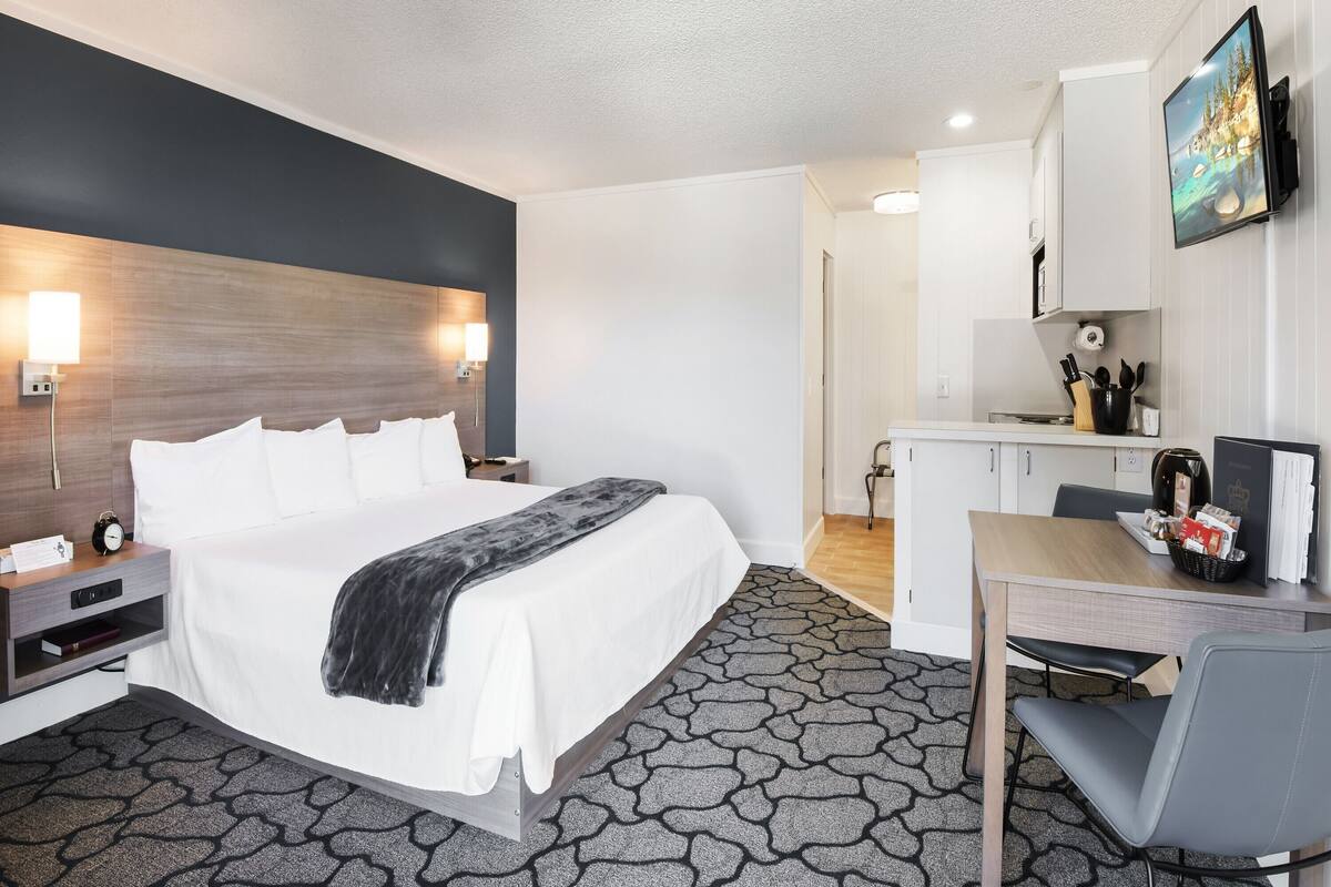 A hotel room with a bed, bedside table, lamp, and a kitchenette area with a TV and cabinets.