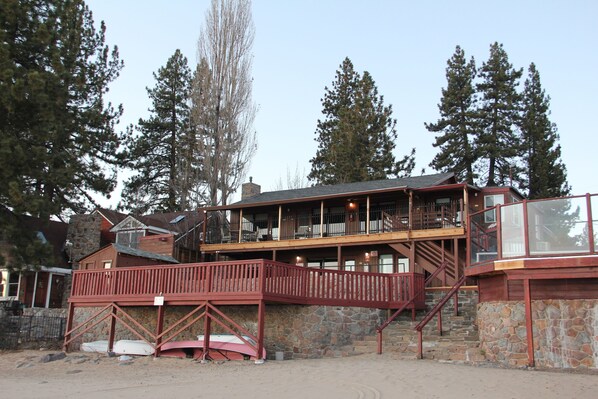 A two-story building with a wooden deck and stone walls, surrounded by tall trees.