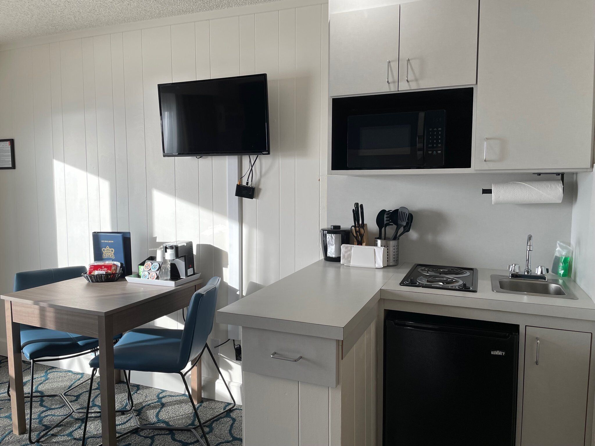 A compact kitchen with a microwave, stove, and dishwasher. There's a TV mounted above the counter and a small dining area with chairs and a table.