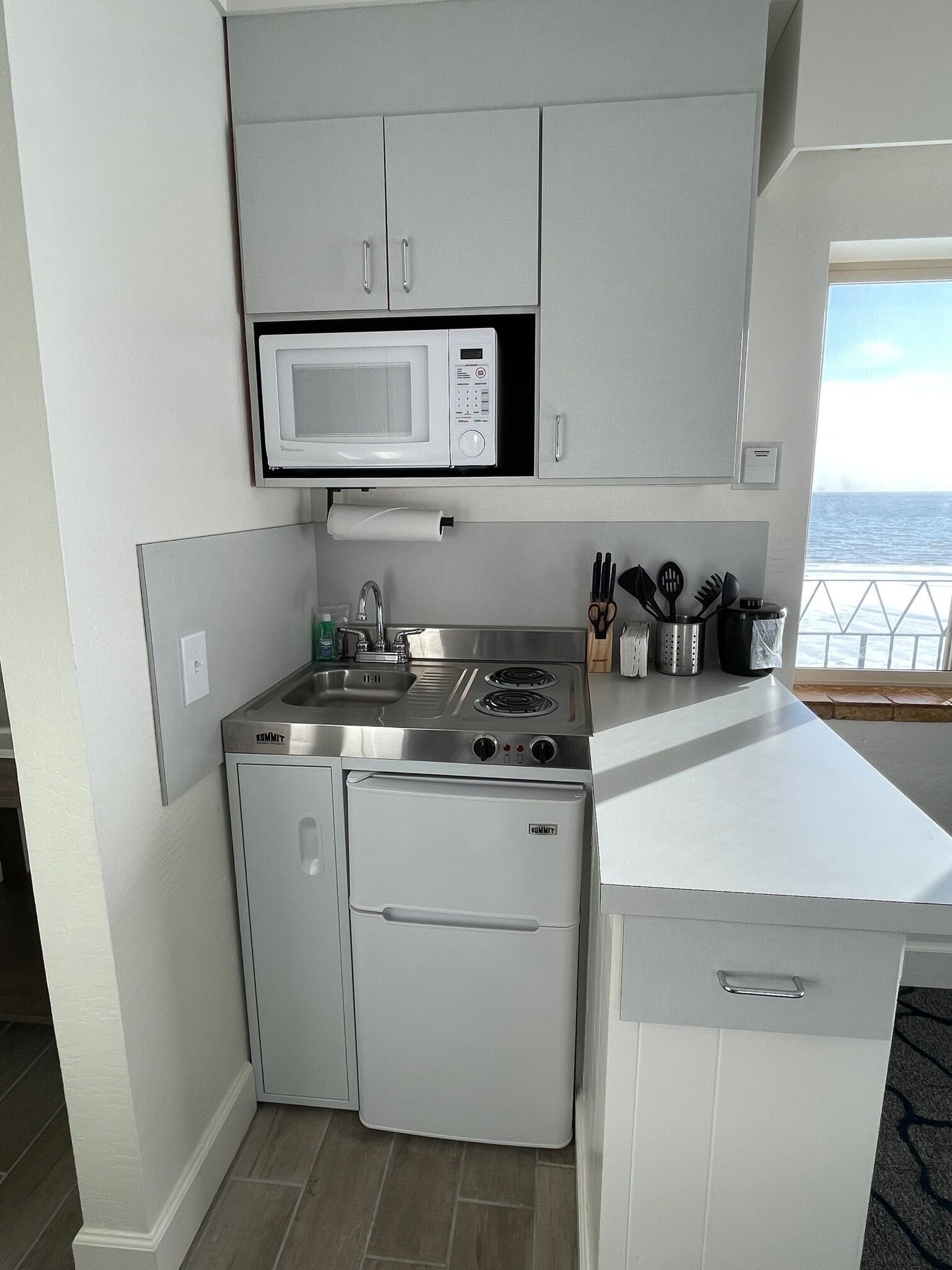A compact kitchen with a microwave, stove, and refrigerator, featuring a view of the sea through a window.