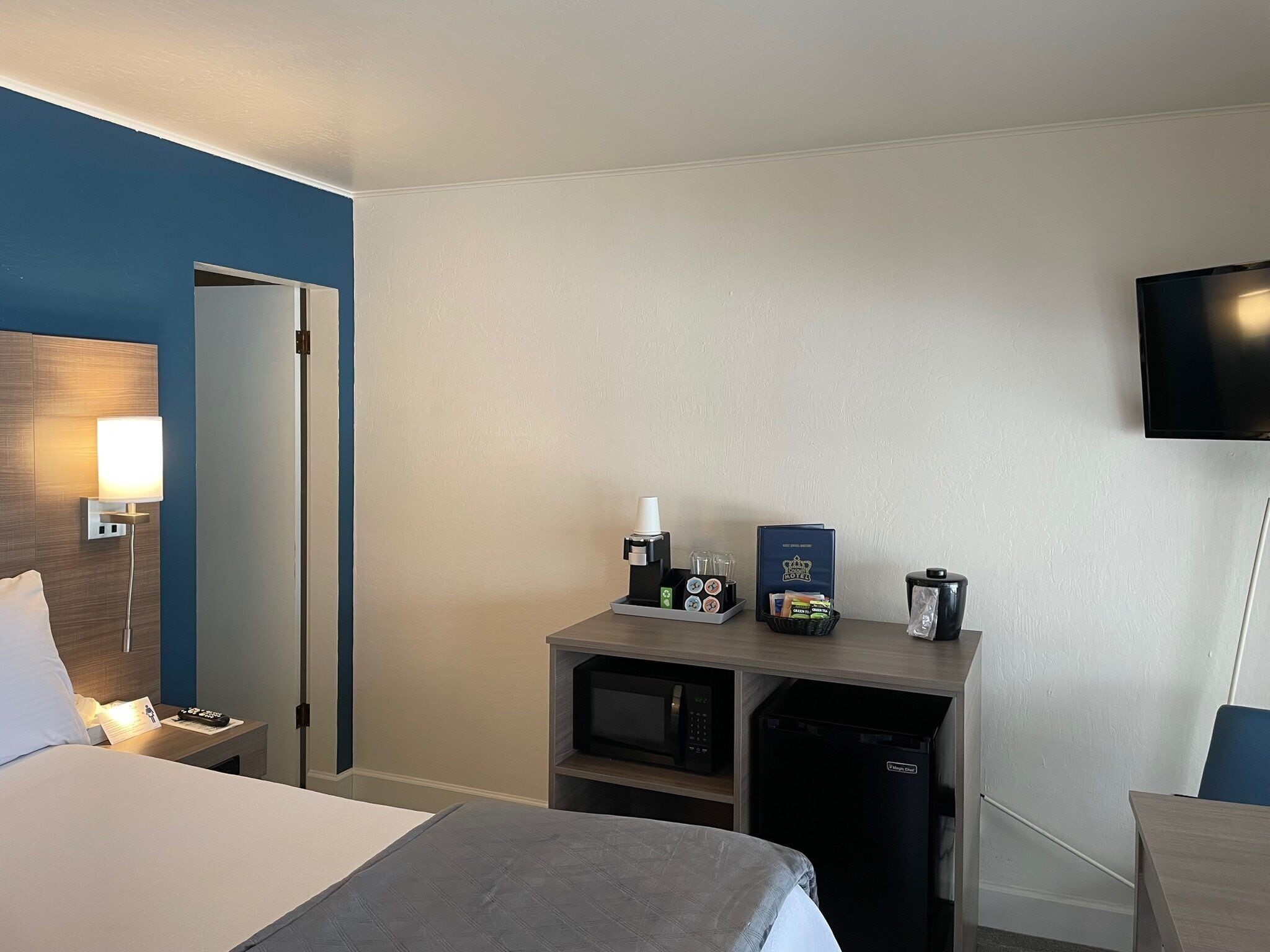 Habitación de hotel con cama, mesita de noche con lámpara, cafetera, microondas, mini refrigerador, televisor y puerta.