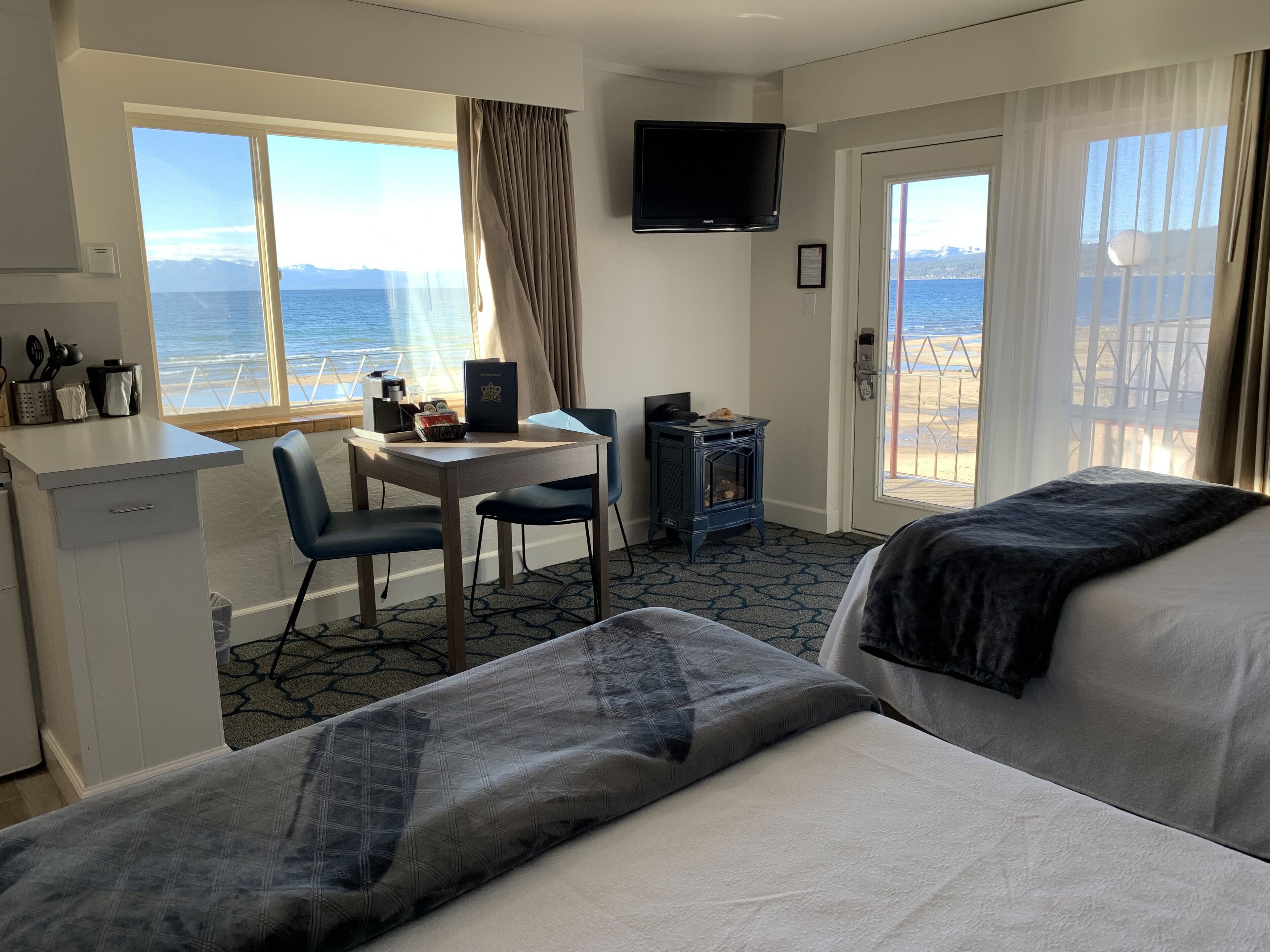 A hotel room with two beds, a desk with a chair, a small table with a book, a television, and a view of the sea.