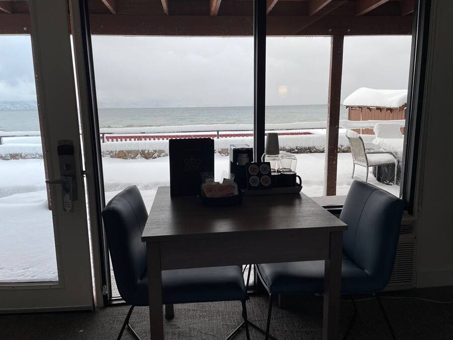 A room with a view of the ocean through large windows, a wooden table, two chairs, and a coffee machine.