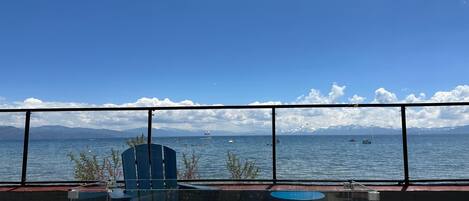A deck with a glass enclosure, a chair, and a view of a lake with boats and mountains in the distance.