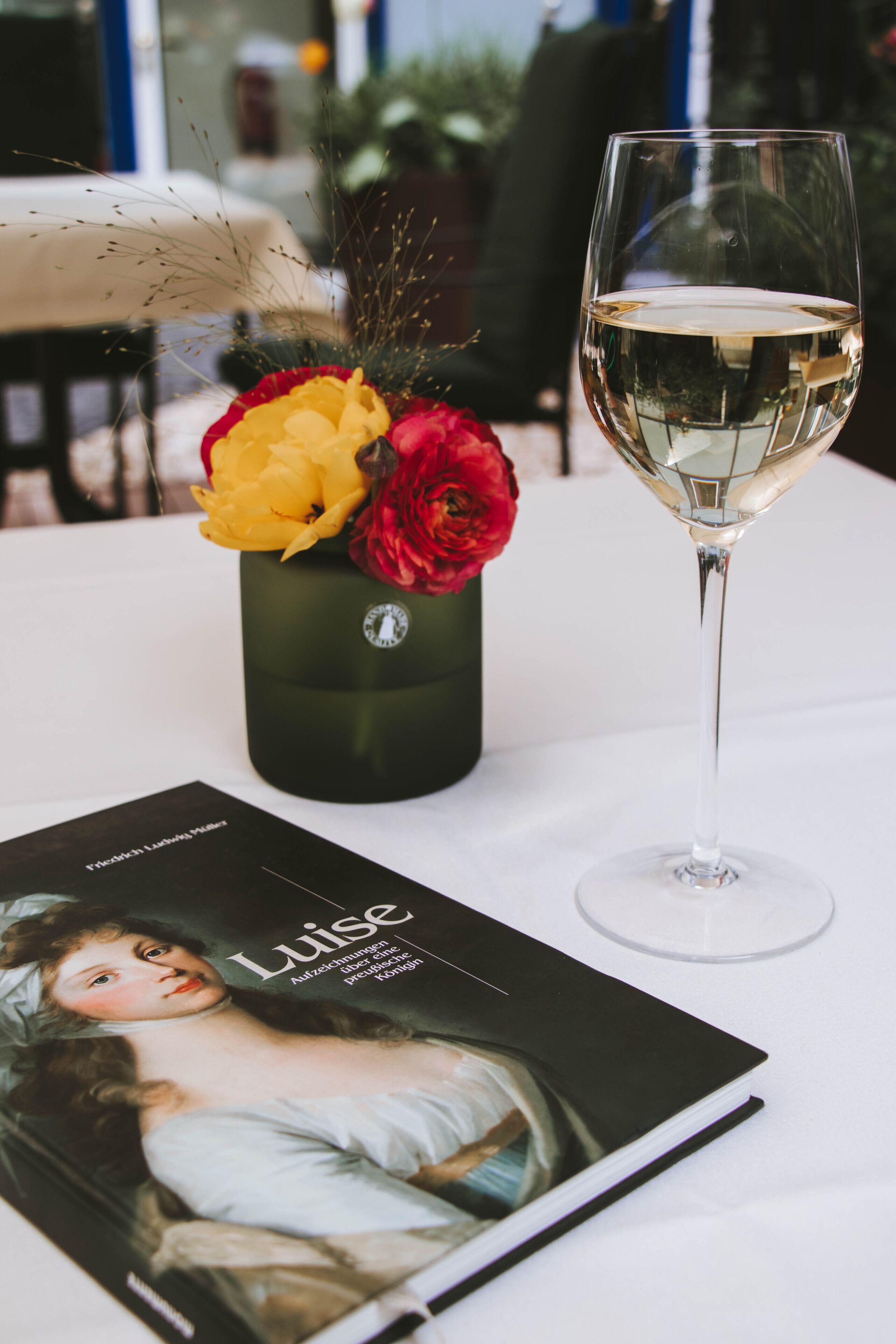A table with a glass of white wine, a black book, and a vase with red and yellow flowers.
