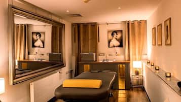 Ett spa-rum med en massagebänk, speglar, en sminkbord med handfat och inramade tavlor på väggen.