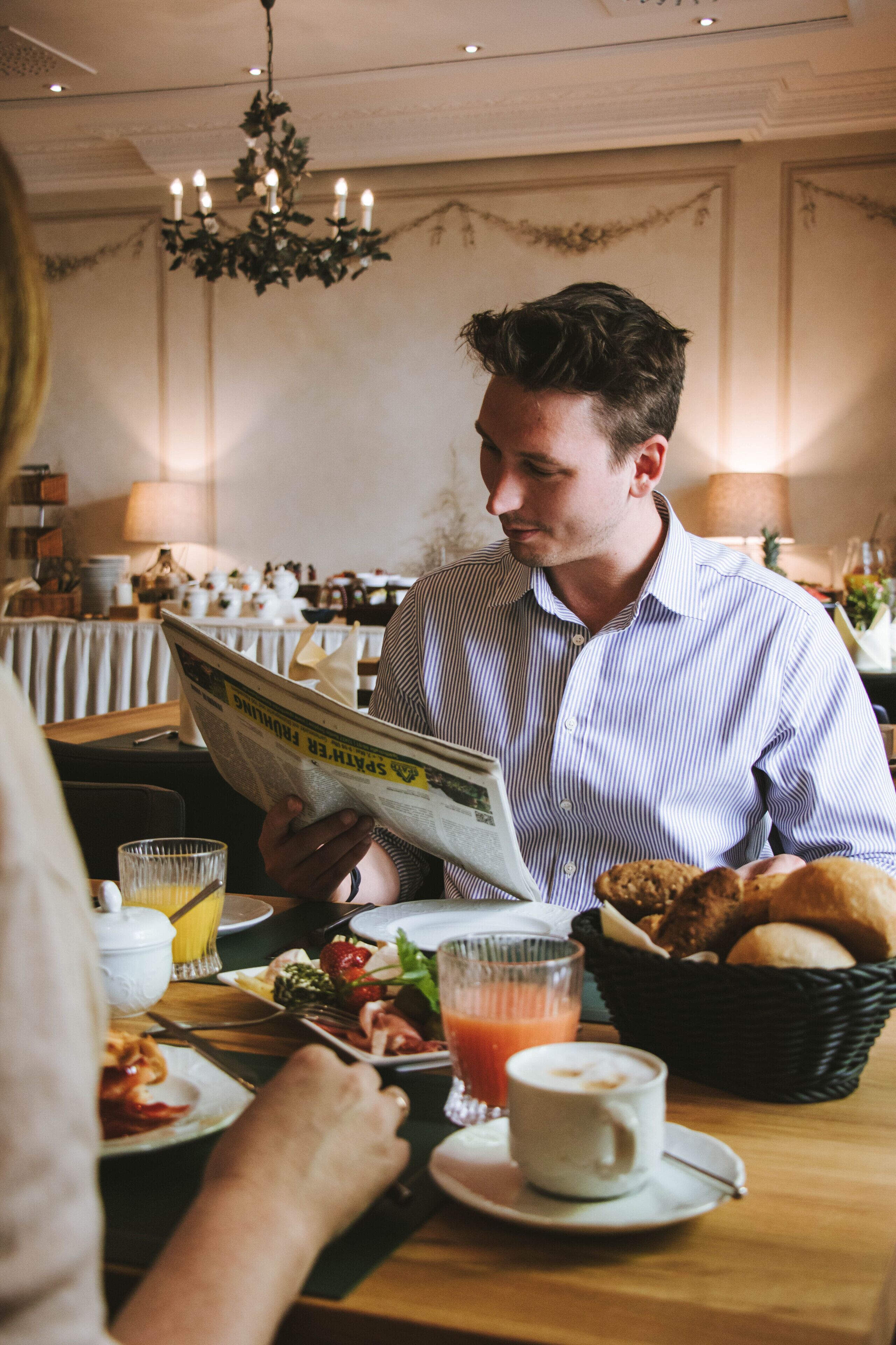 A man reading a newspaper at a dining table with breakfast items, including toast and juice, in a restaurant setting.
