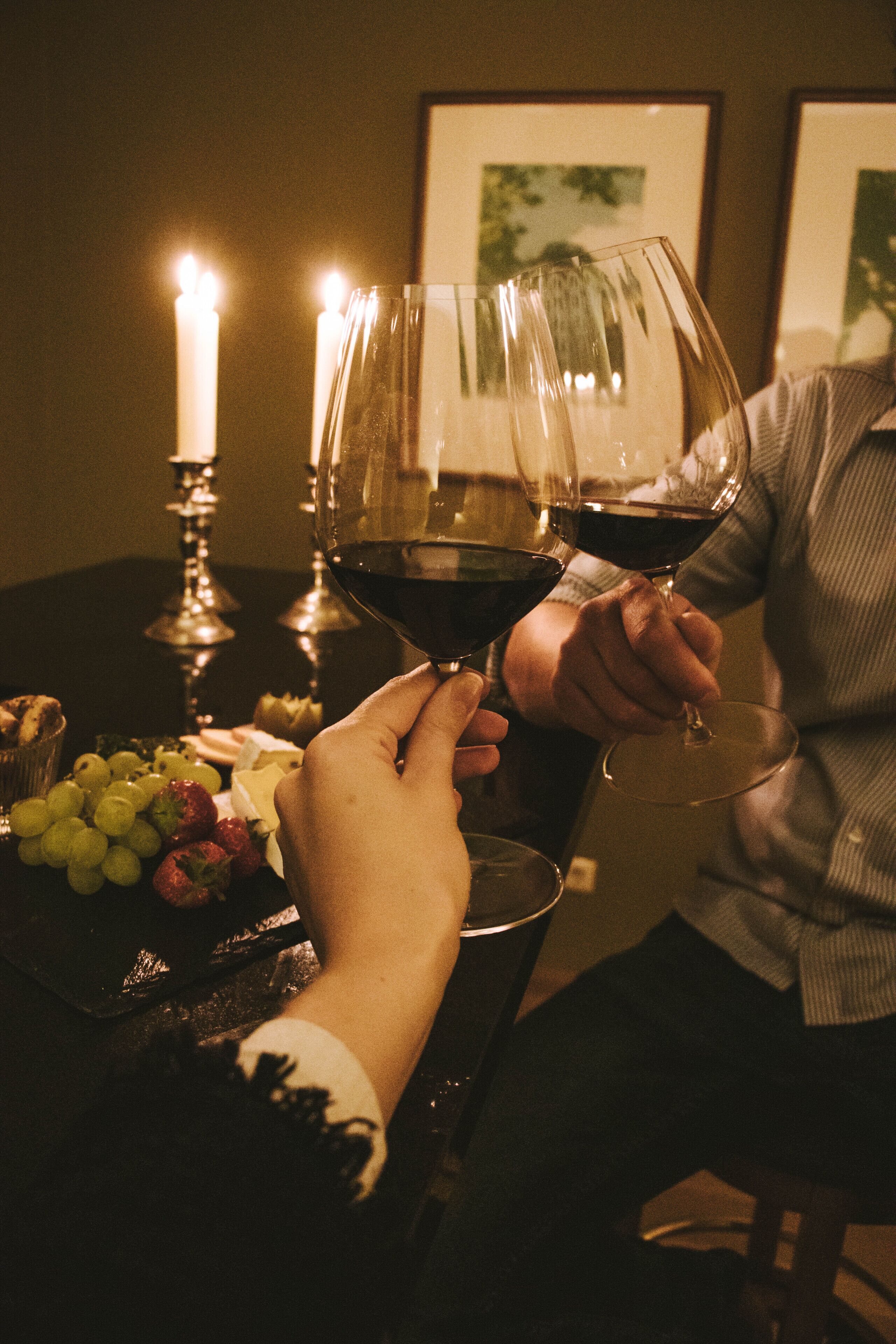 Two people toasting with wine glasses, a candle, and a plate with grapes and strawberries on a table.