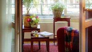 A cozy room with a wooden floor, a red rug, a small table with a cup and a book, a chair with a plaid blanket, and a window with a view of trees and a street.