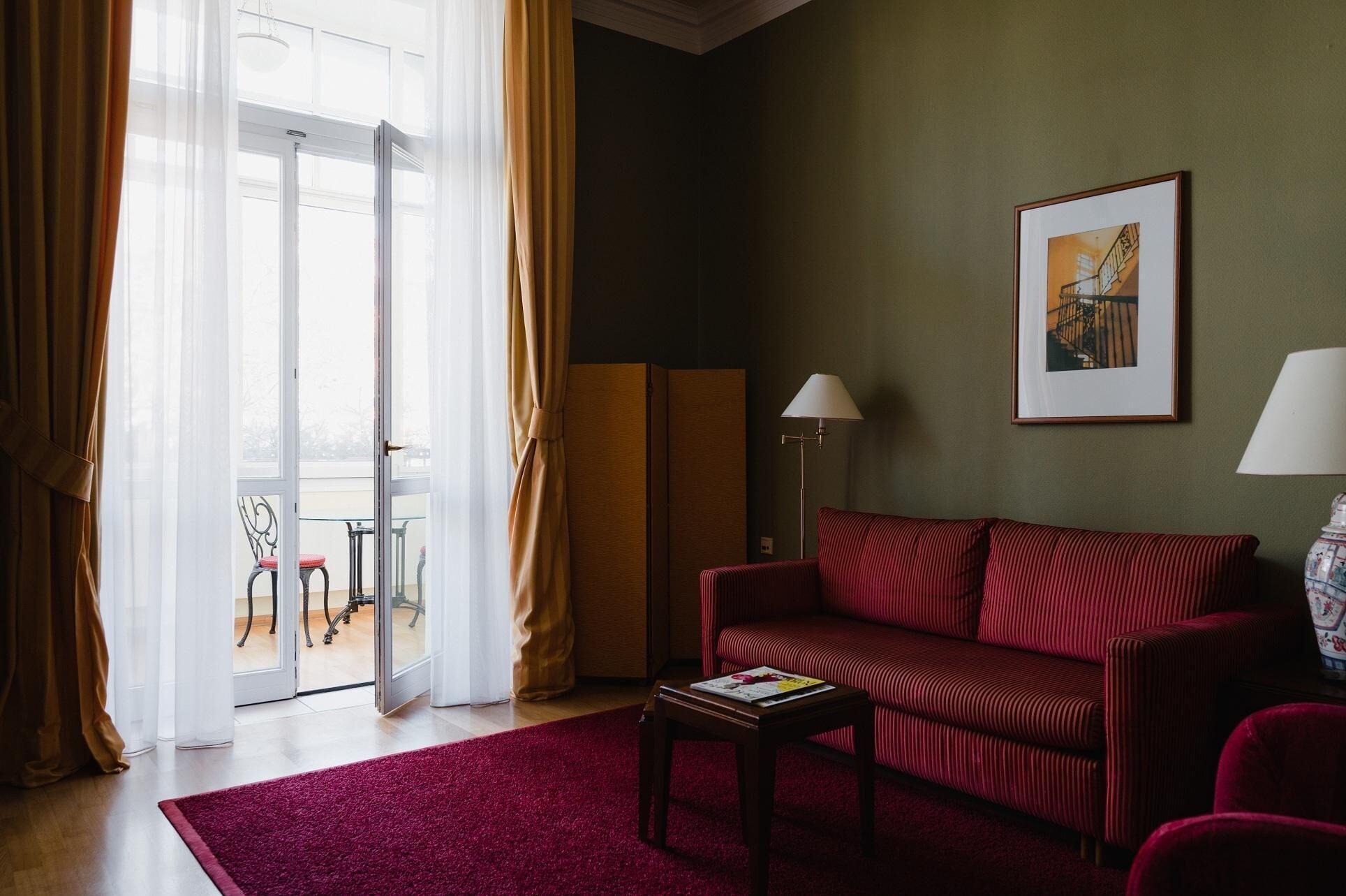 A room with a red sofa, a small table with a magazine, a lamp, a framed picture on the wall, and a view through a glass door.