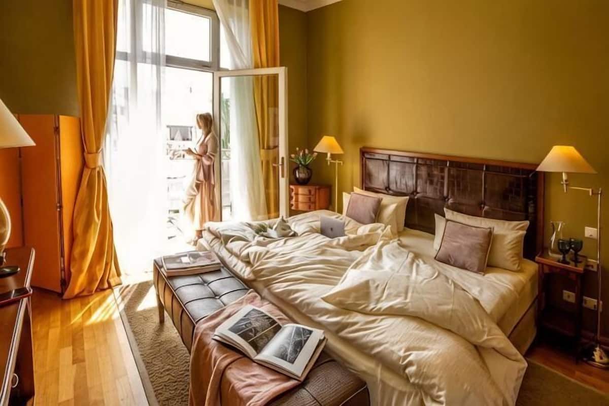 A double bed with a wooden headboard, bedside tables with lamps, a bench with a book, and a large window with curtains.