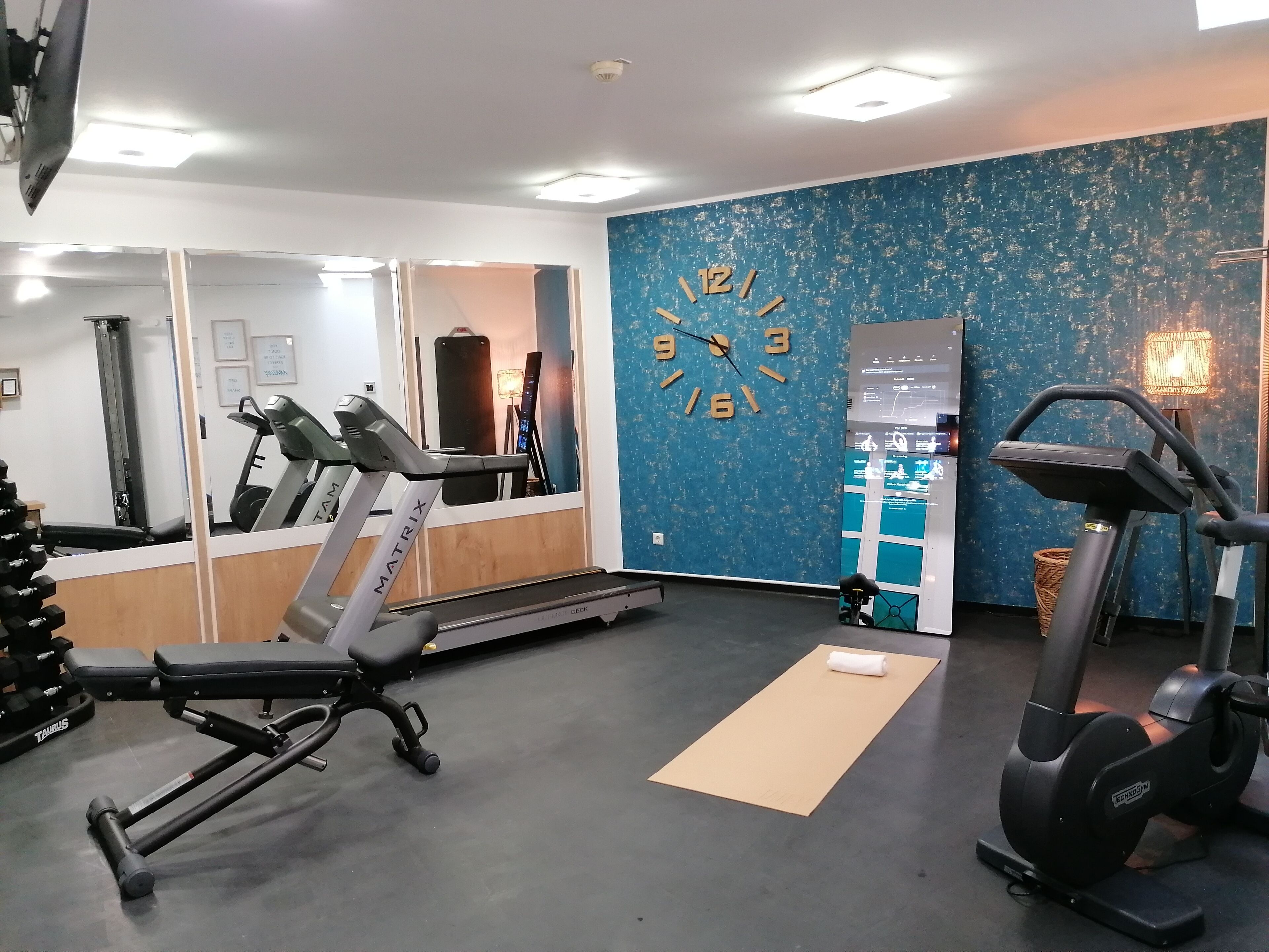 A gym with cardio equipment, a mirrored wall, and a blue textured wall with a clock.