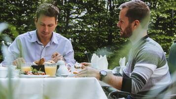 Dos personas cenando en una mesa al aire libre con mantel blanco, rodeadas de follaje.
