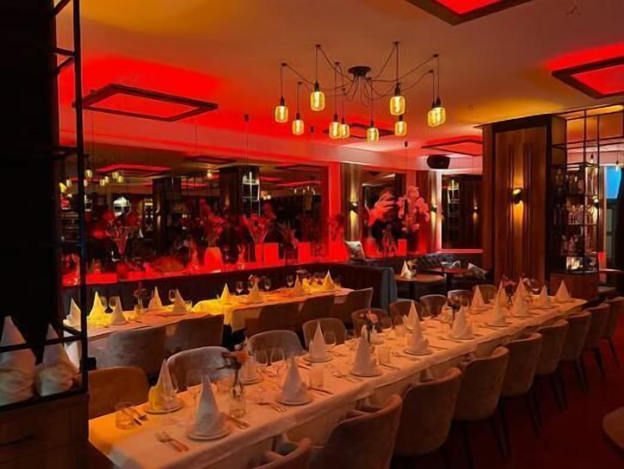 Un restaurante con largas mesas dispuestas para comer, una zona de bar al fondo y una lámpara moderna.