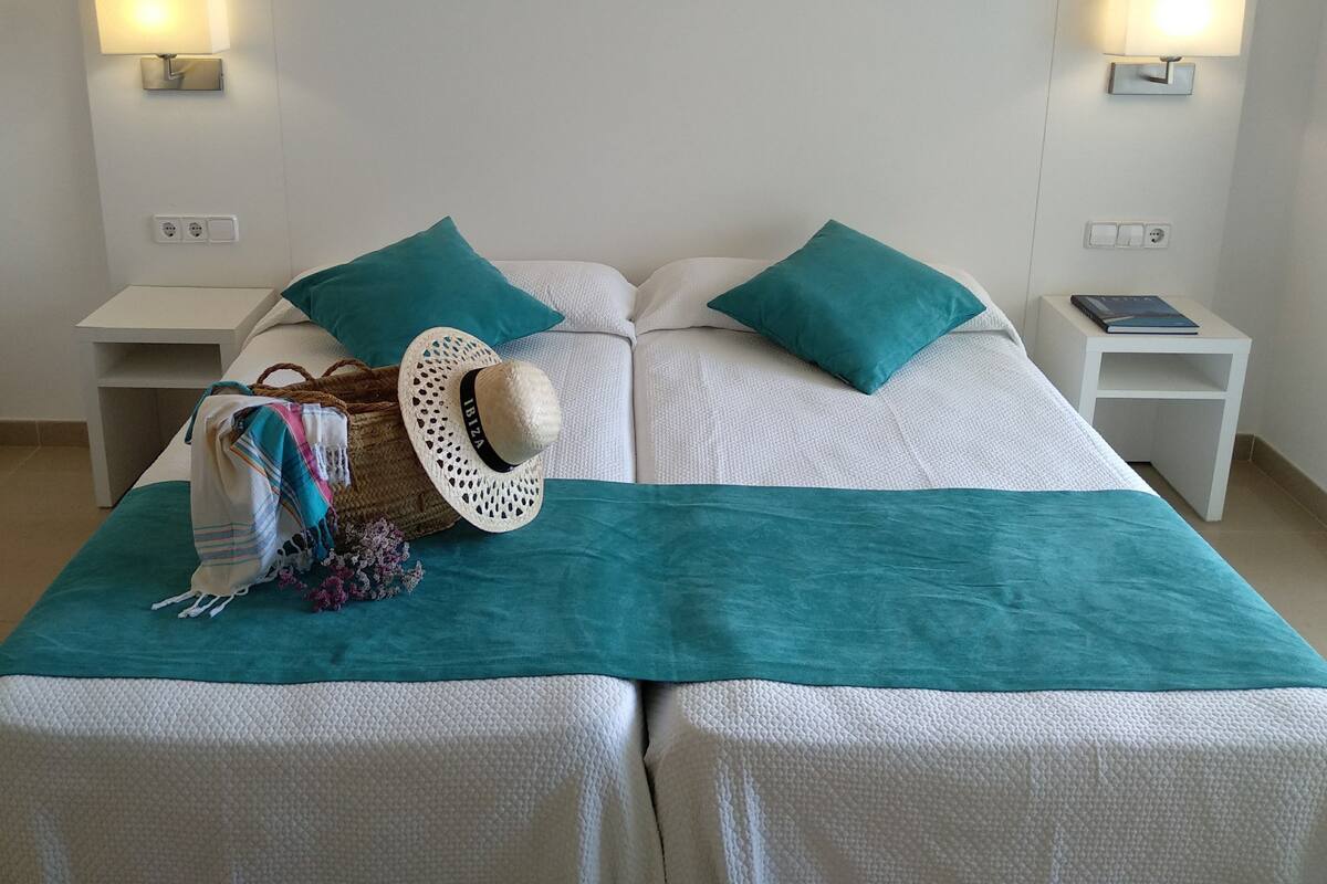A double bed with teal bedding, a white hat, and a woven bag on the bed. There are two white nightstands with lamps, a small table with a book, and a painting of a coastal scene above the bed.