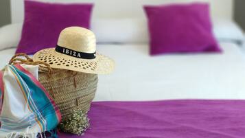 A bed with purple pillows, a straw hat, and a colorful bag on a purple bedspread.