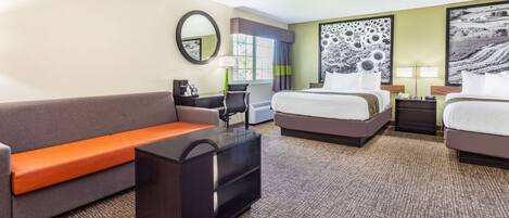 A hotel room with a sofa, two beds, a desk, a mirror, and artwork on the walls.