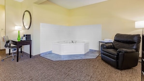Een hotelkamer met een hoekbad, een bureau met een lamp, een lederen fauteuil en een bijzettafeltje met een lamp.