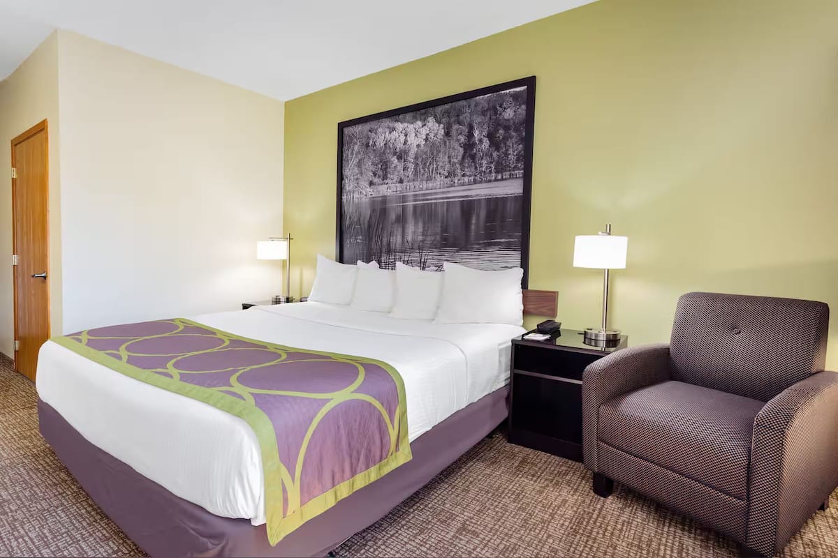 Habitación de hotel con cama, dos mesitas de noche, una silla, un cuadro grande en la pared y una puerta.