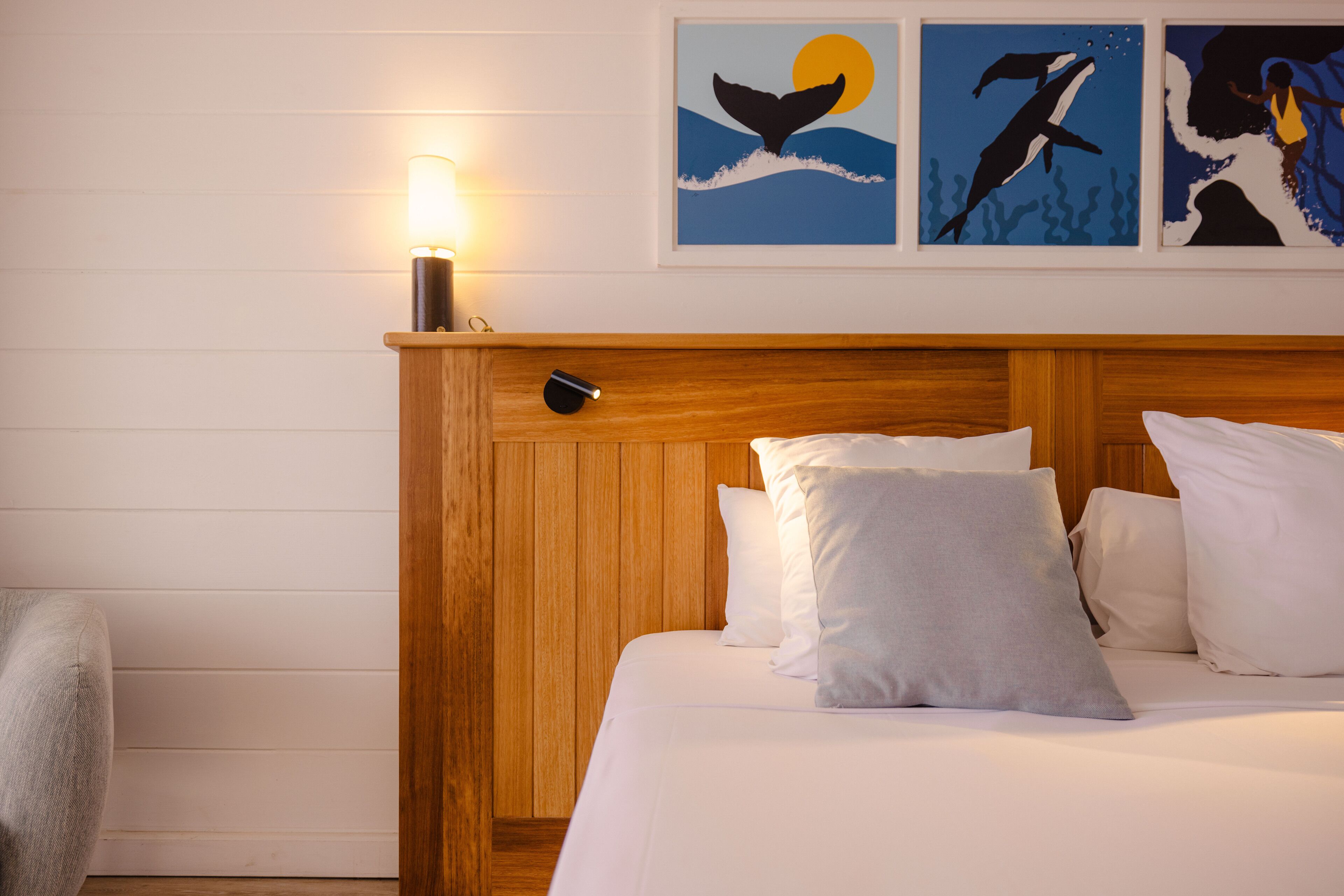 A neatly made bed with white linens and a wooden headboard, a bedside lamp, and three framed artworks depicting whales on a white wall.