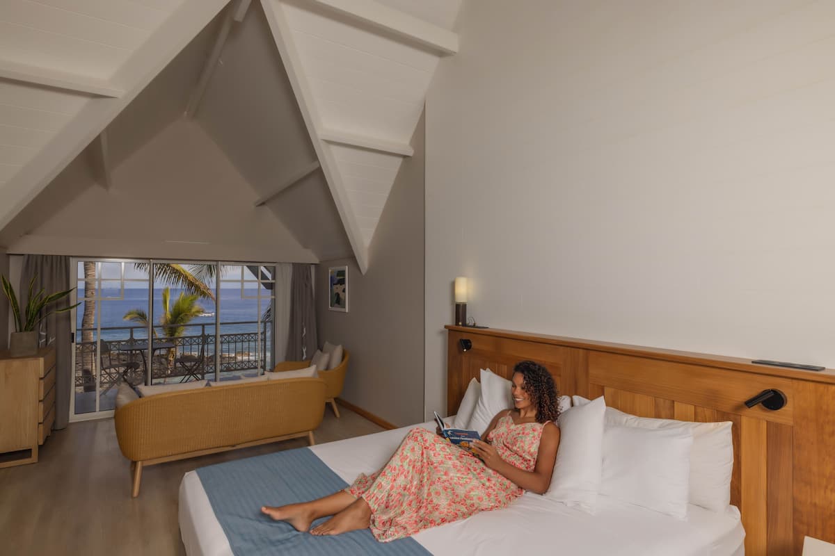 Un dormitorio con cama, mesita de noche, ventana con vista al océano y balcón con una silla y una planta.